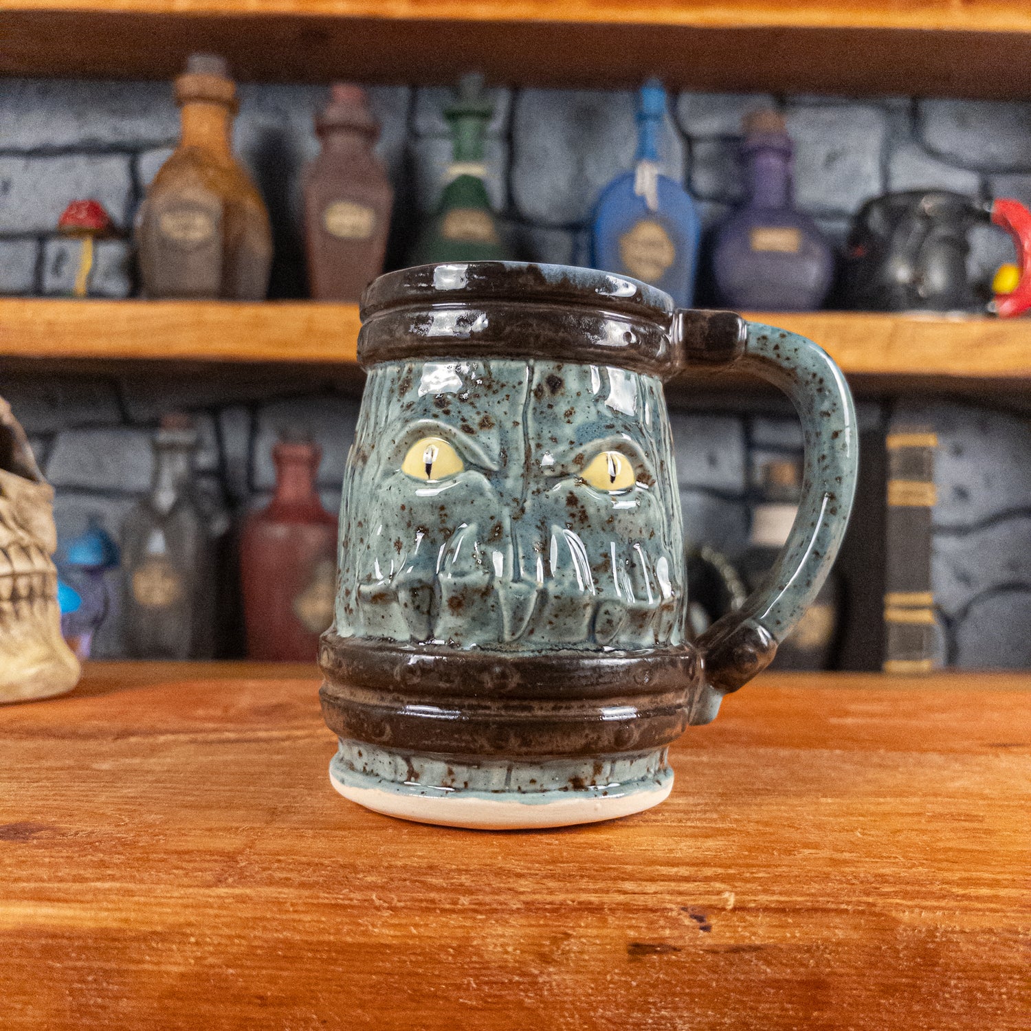 Front view of a handmade Mimic Mug designed to look like a speckled light blue wooden barrel with dark brown bands, featuring menacing yellow eyes and teeth. The mug has a handle.